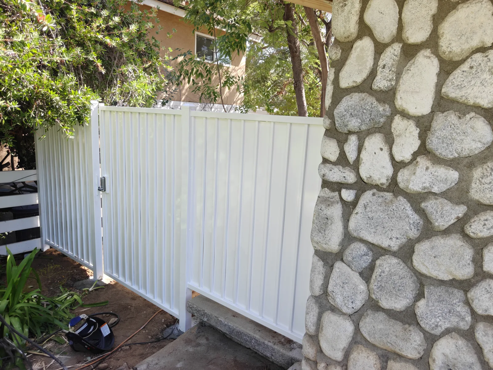 White steel panel privacy fence tied into a river-rock wall at a side entrance — a real Apexguard Fence project in West Covina, CA